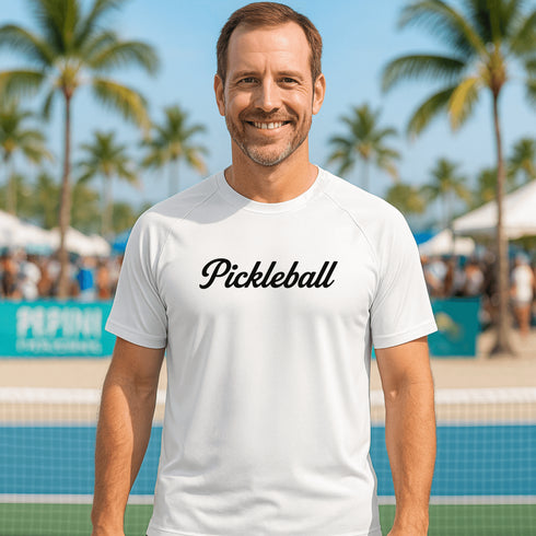 Man wearing a white 'Pickleball' t-shirt on a pickleball court with palm trees in the background.